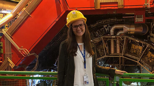 Person smiling in front of a large machine with a hard hat and id tag.