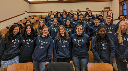 large group of people wearing same hoodie standing in front of chairs and smiling.