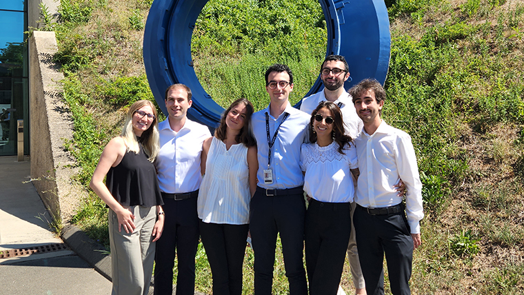 Group of 7 people standing in front of blue circular sculpture in front of building.