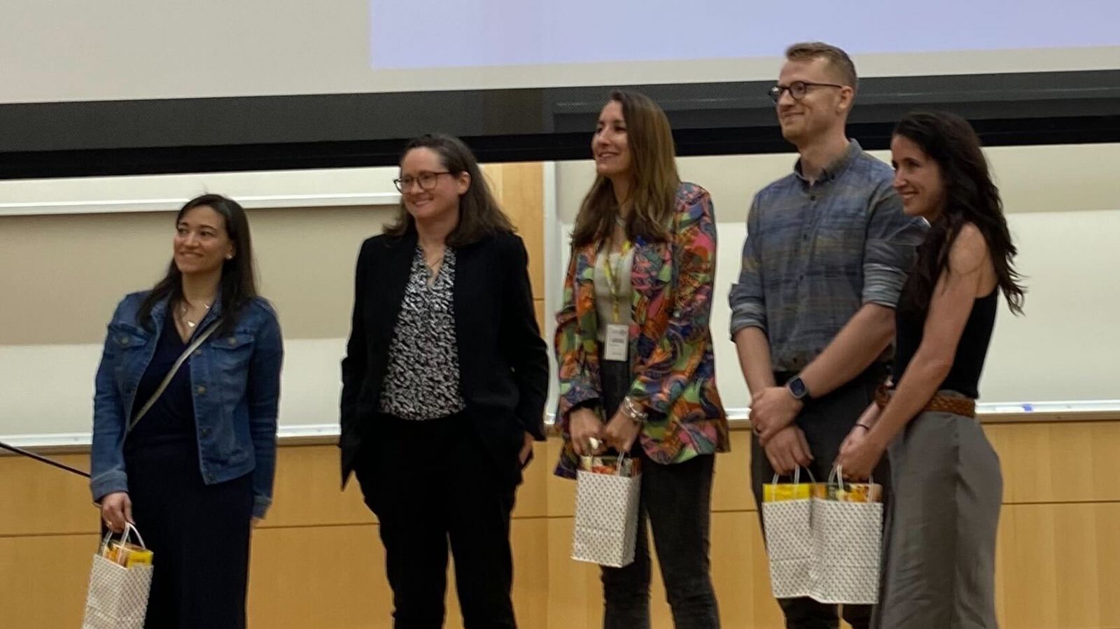 people standing on a stage in front of a screen smiling holding bags.