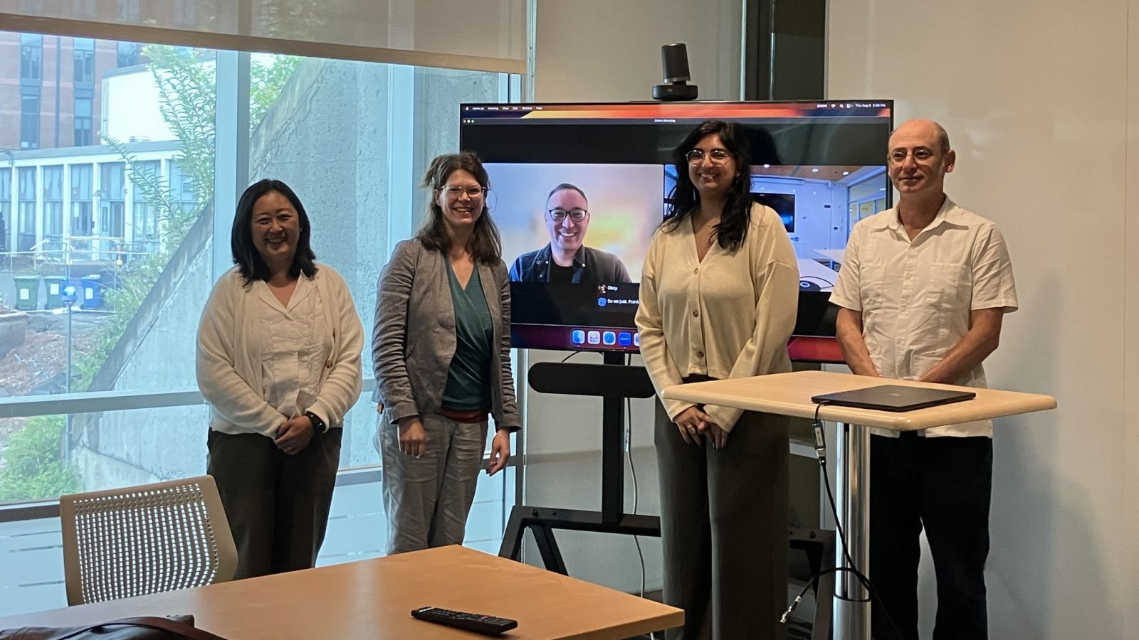 four people standing posing with fifth person in background on zoom.