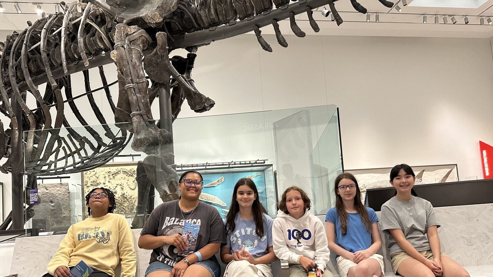 students sitting on a bench posing in front of a large skeleton of dinosaur.