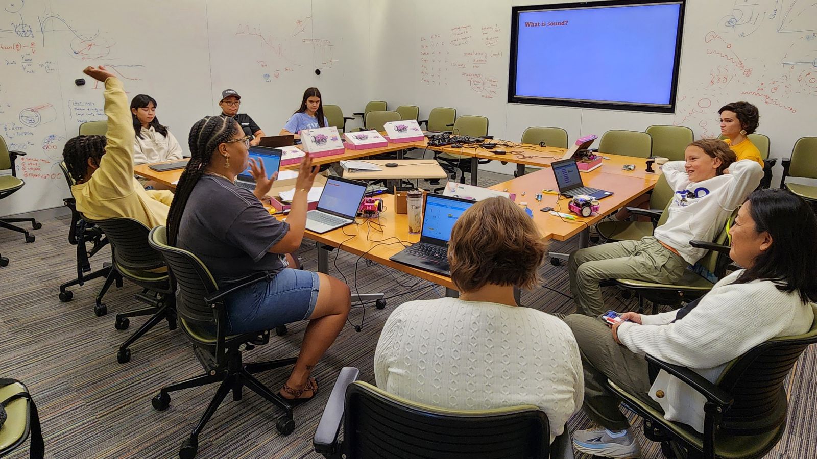 students sitting around a table listening to lecture and looking at screen.