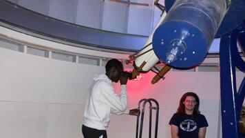 oerson looking through an eyepiece of a large telescope with another person standing in background.