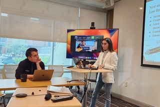 Celin Hidalgo (standing) and Sergio Nuñez (seated) presenting their work for the DOE traineeship in front of a display and a screen.