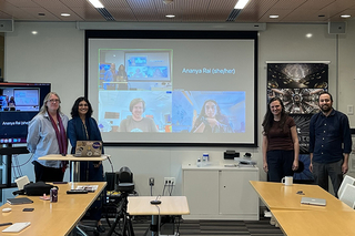 Thesis committee of Ananya Rai standing in room in front of zoom screen with some of the committee on it.