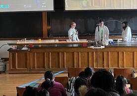 3 people in white lab coats behind a counter with an audience in seats in the foreground.