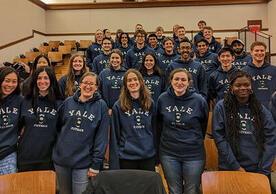 large group of people wearing same hoodie standing in front of chairs and smiling.