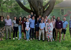 group of people smiling and standing with a fence, tree, and house in the background.