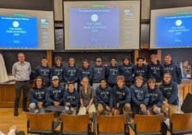 Group of Yale Physics majors in hoodies with two faculty members posing for a picture in a classroom.