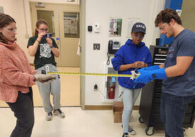 4 people in a lab holding two parts of something with a tape measure in between it.