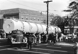 Large tank on truck turning a corner with a group of people watching.