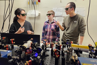 Faculty member mentoring 2 grad students at optical table for the RAY experiment.
