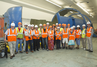 large group of people wearing orange vests standing in front of dismantled blue accelerator tank.