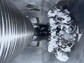 large group of people sitting inside a concrete tube with metal rings overhead.
