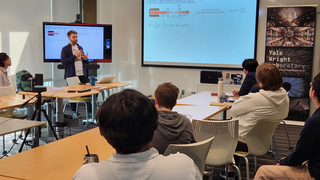Igor Brandao presenting talk in front of screens and audience seated at tables facing speaker.