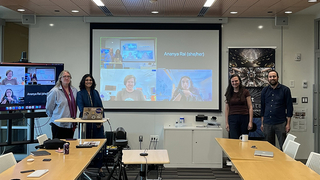 Thesis committee of Ananya Rai standing in room in front of zoom screen with some of the committee on it.