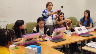 group of students sitting at a table with boxes in front of them listening to instructor who is standing holding a robot.