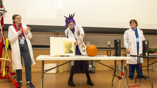 Three people dressed in lab coats doing physics demos at a table with a pumpkin on it.