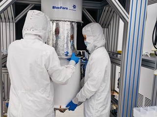 Vasilisa Malenkiy and colleague working in clean room garb on dilution fridge with mirror.