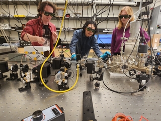 3 QuIPS team members at Wright Lab looking at optical table setup with safety glasses on.