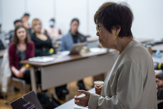 Dr. Keiko Ogura in foreground with Talia Weiss and others at desks listening in backgfound.