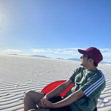 person sitting in a desert scene with sand with waves in it, looking at the sun.