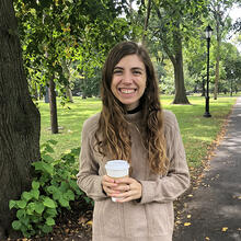 person standing holding coffee cup in front of trees, grass and road.