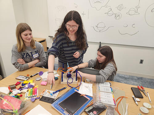 3 people working with art supplies at a table in front of a white board.