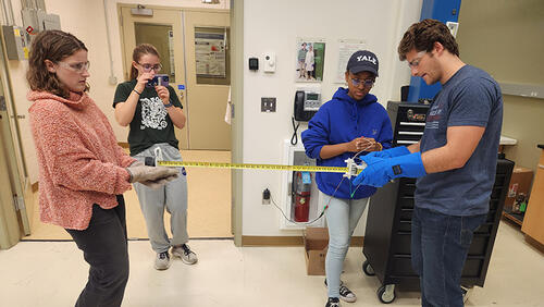 4 people in a lab holding two parts of something with a tape measure in between it.